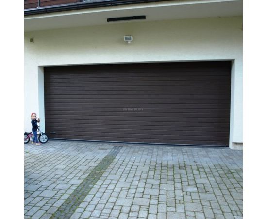 Tumši brūni sekciju garāžu vārti ar dabīgu koka imitāciju un horizontālu struktūru bruģētā pagalma priekšā privātmājai.